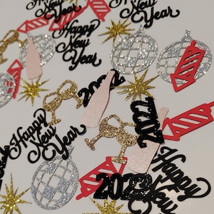 May include: A collection of New Year's Eve decorations scattered across a white surface. The decorations include glittery gold stars, silver disco ball ornaments, and red and black cutouts of fireworks and the text "Happy New Year" and "2022".