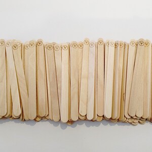 May include: A pile of light-coloured wooden craft sticks, each with a rounded top and a small circular logo. The sticks are arranged closely together, creating a textured surface. The natural wood grain is visible.