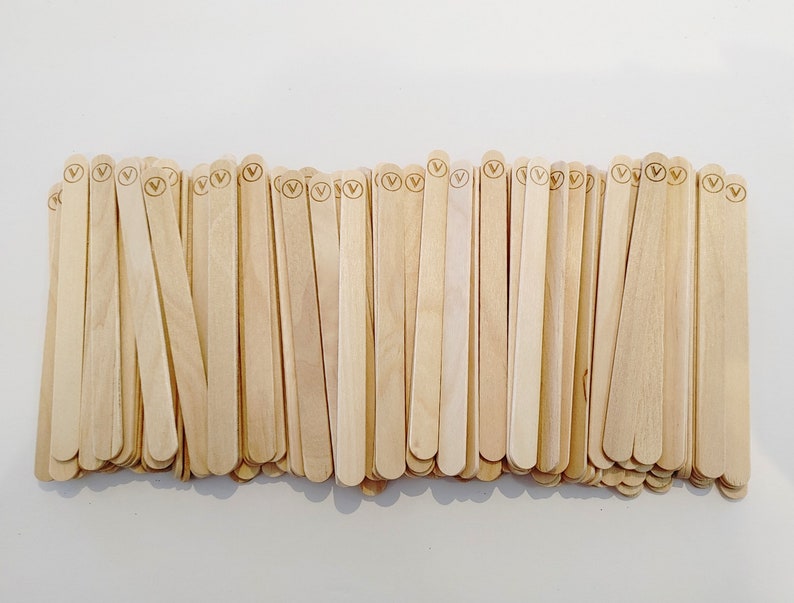 May include: A pile of light-coloured wooden craft sticks, each with a rounded top and a small circular logo. The sticks are arranged closely together, creating a textured surface. The natural wood grain is visible.