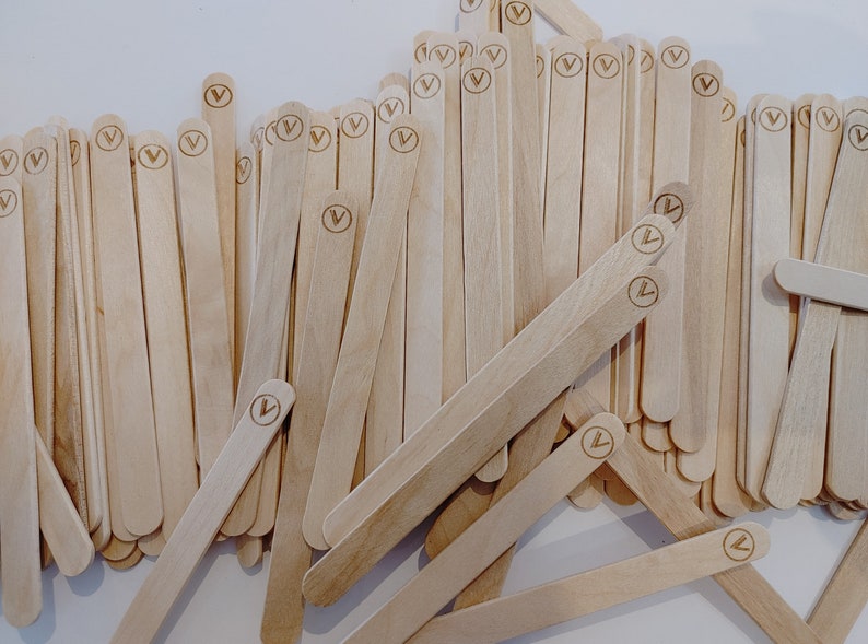 May include: A pile of light-coloured wooden craft sticks, each with a rounded end and a circular logo with a 'V' inside. The sticks are scattered, suggesting a bulk supply for crafts or DIY projects.