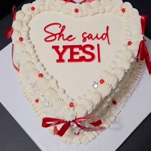 Puede incluir: Un pastel en forma de corazón con glaseado blanco liso, decorado con bordes blancos, pequeños acentos esféricos rojos y lazos de cinta roja. La parte superior del pastel presenta el texto rojo "She said YES" en letras cursivas y de molde.