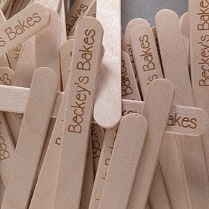 May include: A collection of light-coloured wooden craft sticks, each engraved with the text "Beckey's Bakes". The sticks are smooth with rounded ends, likely for crafts or baking purposes.