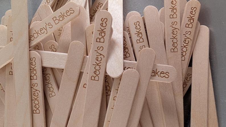 May include: A collection of light-coloured wooden craft sticks, each engraved with the text "Beckey's Bakes". The sticks are smooth with rounded ends, likely for crafts or baking purposes.