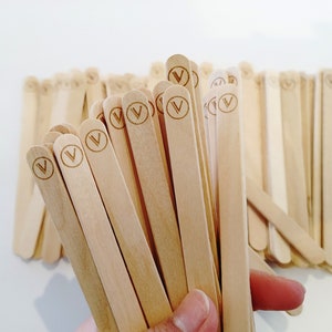 May include: A bundle of light-coloured wooden craft sticks, each with a circular logo containing a stylized "V" etched near the top. The sticks are held together, with some fanned out, against a white background.