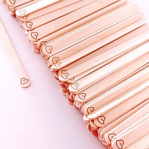 May include: A collection of light-coloured wooden craft sticks, each with a small, smiling face design etched on one end. The sticks are arranged in a neat, angled stack, suggesting a craft or DIY project.