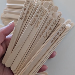 Puede incluir: Un conjunto de palitos de madera claros, cada uno grabado con los nombres "Austin" y "Abby" separados por un corazón. Los palitos se sostienen juntos en un grupo, con otros dispersos en el fondo.