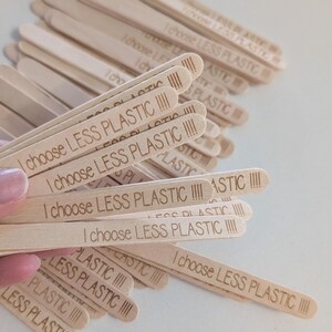 May include: A pile of light-coloured wooden craft sticks, each engraved with the phrase "I choose LESS PLASTIC" in a sans-serif font. The sticks are arranged in a pile, with some held in a hand. The text is clear and legible.