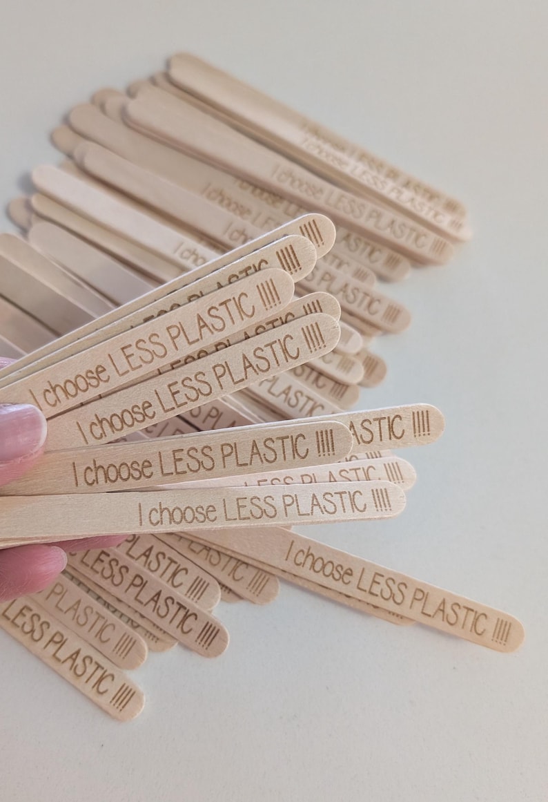 May include: A pile of light-coloured wooden craft sticks, each engraved with the phrase "I choose LESS PLASTIC" in a sans-serif font. The sticks are arranged in a pile, with some held in a hand. The text is clear and legible.