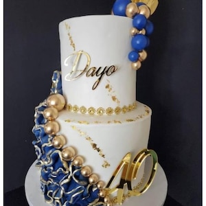 Può includere: Una torta bianca a due piani decorata con accenti in foglia d'oro, un topper dorato con la scritta Dayo e il numero 40 in oro. La torta presenta una cascata di sfere blu e dorate, un bordo floreale dorato e dettagli a balze di colore blu.