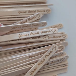 May include: A pile of light-coloured wooden stir sticks, each engraved with the text "Giana's Bridal Shower" and a heart design. The sticks are arranged in a pile, suitable for a bridal shower.