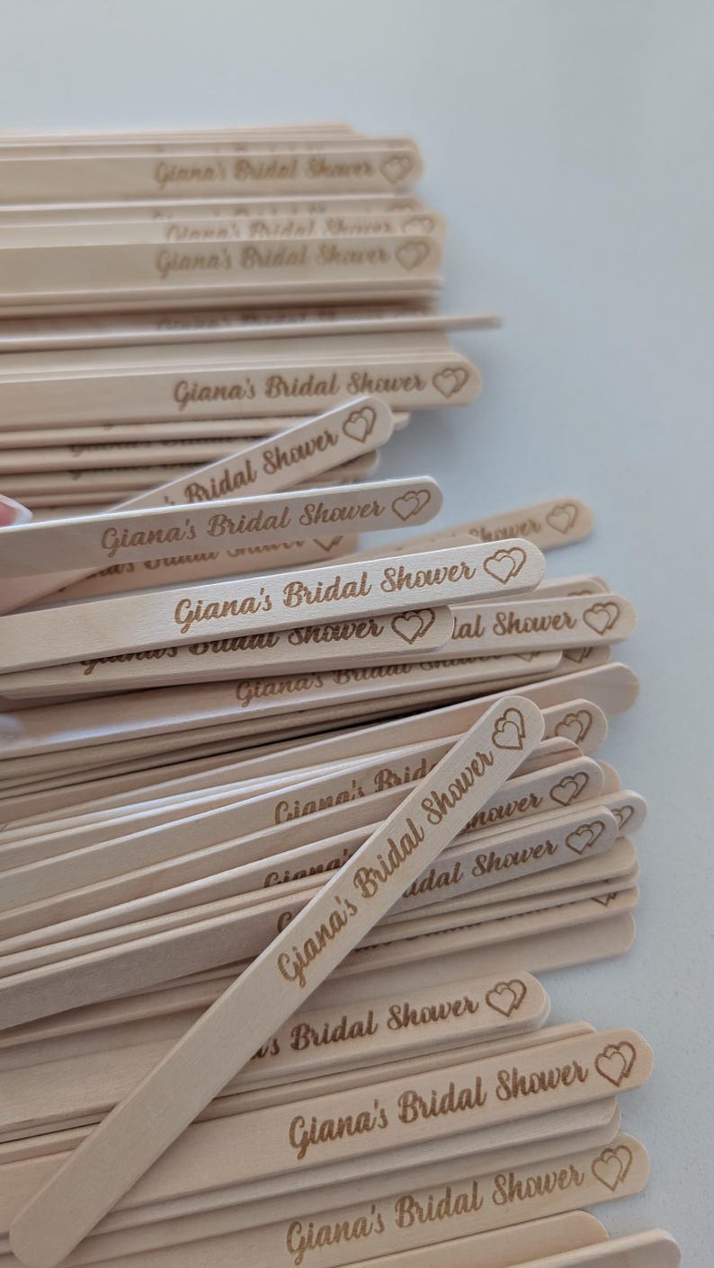 May include: A pile of light-coloured wooden stir sticks, each engraved with the text "Giana's Bridal Shower" and a heart design. The sticks are arranged in a pile, suitable for a bridal shower.