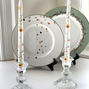Könnte beinhalten: Zwei klare Glas-Kerzenhalter mit floralen Musterkerzen. Die Kerzen sind weiß mit einem floralen Muster in Rosa-, Gelb- und Grüntönen. Die Kerzenhalter stehen auf einer weißen Oberfläche mit einem weißen und grünen Teller dahinter.