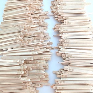 May include: Two stacks of wooden craft sticks are shown. Each light brown stick has the word "CALLAB" printed on it. The sticks are arranged in neat piles, suitable for craft projects. The image is well-lit.