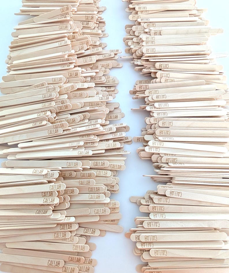 May include: Two stacks of wooden craft sticks are shown. Each light brown stick has the word "CALLAB" printed on it. The sticks are arranged in neat piles, suitable for craft projects. The image is well-lit.