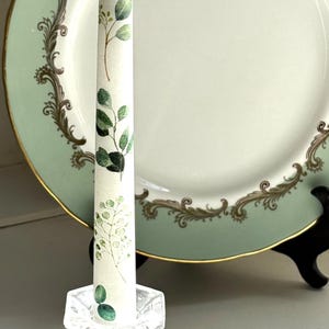 Puede incluir: Una vela blanca con un diseño floral verde se encuentra sobre un candelero de vidrio transparente. La vela está sobre una superficie blanca con un plato decorativo verde y dorado detrás.