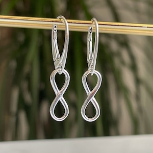 May include: A pair of silver infinity symbol earrings with a delicate design. The earrings are hanging from a gold bar.