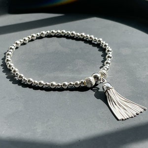 May include: A silver beaded bracelet with a silver tassel charm. The bracelet is made of small, round beads and has a simple, elegant design.