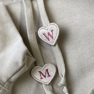 Puede incluir: Una sudadera con capucha de color beige que presenta dos cordones planos tejidos. Cada cordón está adornado con un parche en forma de corazón bordado con una letra rosa oscuro, las letras W y M, centradas sobre la tela de la sudadera.