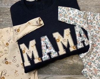 May include: A black t-shirt with the word "MAMA" spelled out in large letters. Each letter is made of a different floral fabric. The t-shirt is paired with a matching baby onesie.