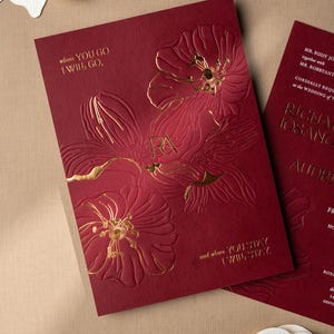 May include: Burgundy wedding invitations with gold floral embossing and the initials "RA". The text "where YOU GO I WILL GO" and "and where YOU STAY I WILL STAY" are printed on the invitation. White floral cutouts are in the background.
