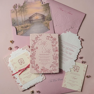 May include: A wedding invitation suite featuring textured paper cards with pink floral illustrations, ornate die-cut edges, and a pink envelope with a printed landscape scene. The text includes names like Randhawa and The Brar Family.