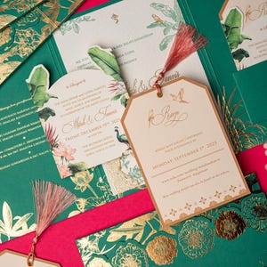Può includere: Elegante suite di inviti di nozze in verde smeraldo e oro, con disegni floreali e botanici. Include biglietti d'invito, tag RSVP con nappe e buste decorative. Il set è ambientato su uno sfondo rosa acceso.