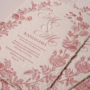 May include: Off-white textured paper wedding invitation with deckled edges, featuring a red floral border and embossed text: Mr & Mrs RANDHAWA, joyfully invite you to celebrate their love and marriage, Friday, the eighth of August, two thousand twenty-five, at five thirty in the evening, Grand Empire Banquet Hall, 12888 80 Ave Unit 207, Surrey, BC.