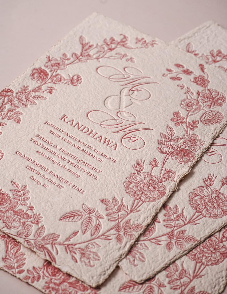 May include: Off-white textured paper wedding invitation with deckled edges, featuring a red floral border and embossed text: Mr & Mrs RANDHAWA, joyfully invite you to celebrate their love and marriage, Friday, the eighth of August, two thousand twenty-five, at five thirty in the evening, Grand Empire Banquet Hall, 12888 80 Ave Unit 207, Surrey, BC.
