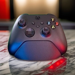 Peut inclure: Une manette Xbox noire avec des lumières bleues et rouges sur le dessous. La manette a un motif en damier noir et blanc sur le devant.