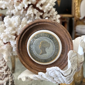 May include: A vintage Cherry Tooth Paste tin in a wooden holder. The tin features a profile of a woman and text that reads "Prepared by John Gosnell & Co London". The wooden holder is round and has a dark brown finish.