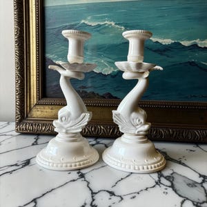 May include: A pair of white ceramic candle holders, each featuring a sculpted dolphin base with an upward-curving tail that supports a fluted candle cup. The circular bases are decorated with a subtle embossed pattern along the bottom edge.