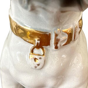 May include: A ceramic pug figurine with a gold-colored collar and clasp. The pug has a white body with black and grey markings on its head. The collar is detailed with gold paint.