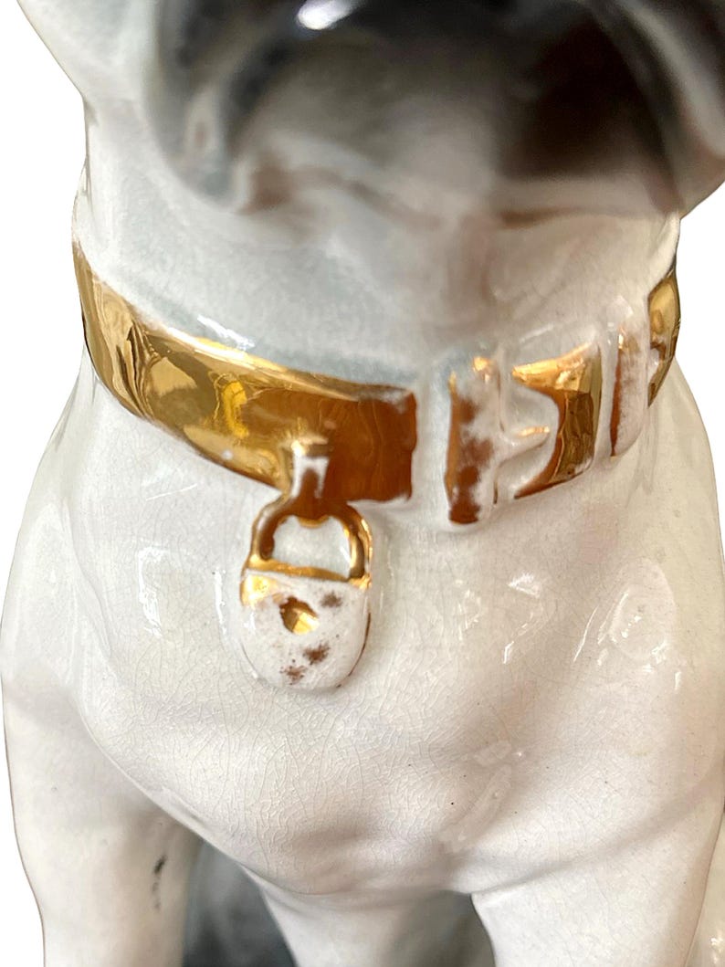 May include: A ceramic pug figurine with a gold-colored collar and clasp. The pug has a white body with black and grey markings on its head. The collar is detailed with gold paint.