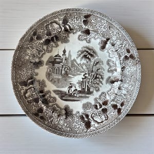 May include: A brown and white ceramic plate with a floral and landscape design. The plate features a detailed illustration of a building and figures, surrounded by a floral border. The rim has a decorative pattern.
