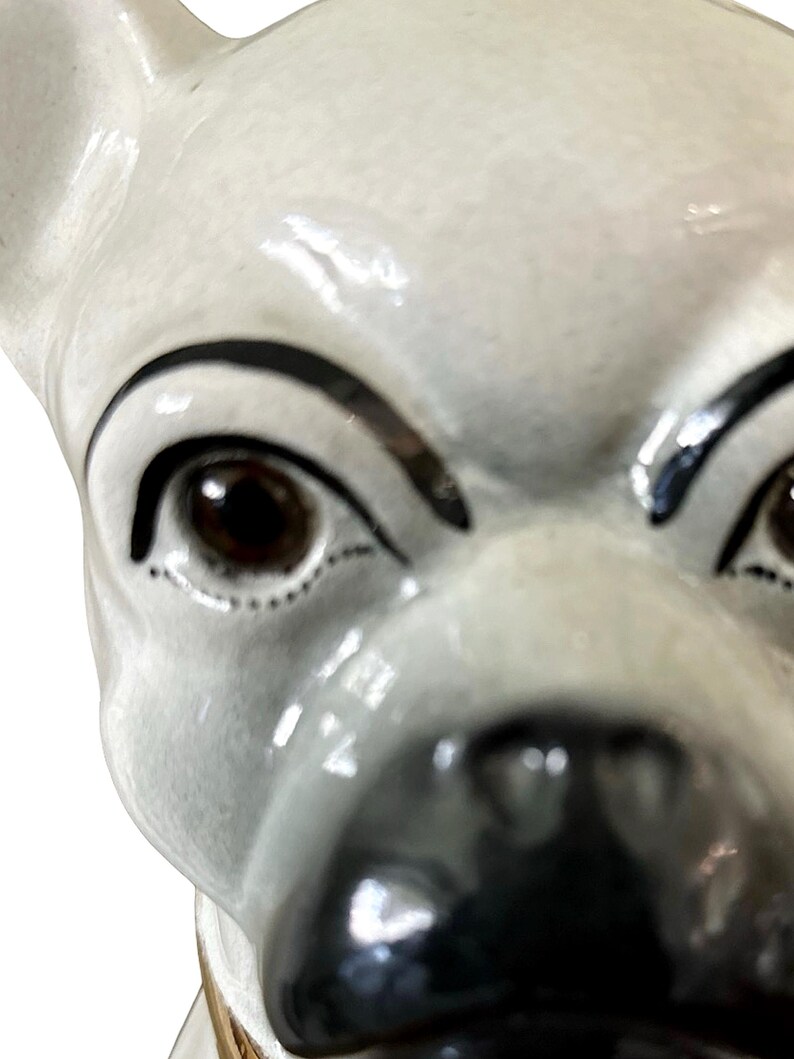 May include: A close-up of a ceramic dog figurine. The dog has a white body with black accents around the eyes and nose. The eyes are brown and the figurine has a glossy finish.