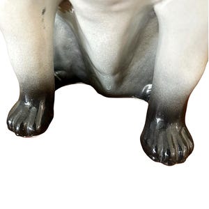 May include: A ceramic figurine of a dog, viewed from the underside. The dog's legs and paws are visible, with a gradient of gray to black coloring. The figurine has a glossy finish and is set against a white background.