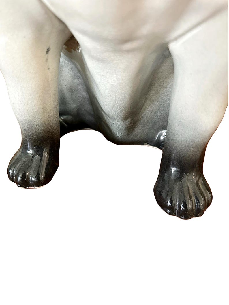 May include: A ceramic figurine of a dog, viewed from the underside. The dog's legs and paws are visible, with a gradient of gray to black coloring. The figurine has a glossy finish and is set against a white background.