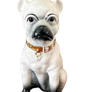 May include: A ceramic bulldog figurine in a seated position. The dog is primarily white with black accents on the nose, paws, and ears. It wears a gold-colored collar with a small tag. The figurine has a stern expression.