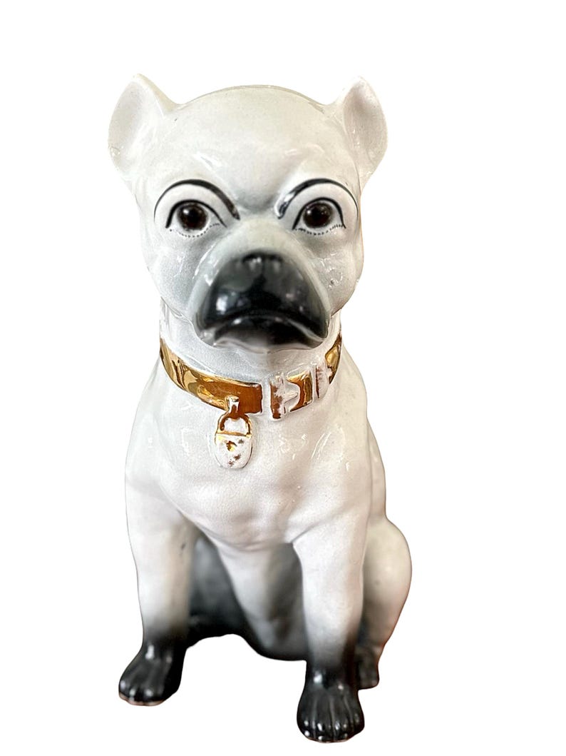 May include: A ceramic bulldog figurine in a seated position. The dog is primarily white with black accents on the nose, paws, and ears. It wears a gold-colored collar with a small tag. The figurine has a stern expression.
