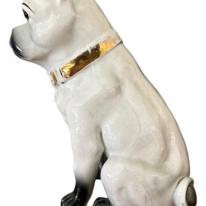 May include: A ceramic dog figurine in a sitting position. The dog is primarily white with black accents on the paws, nose, and ears. It has a gold-colored collar. The figurine has a glossy finish and is likely a decorative item.