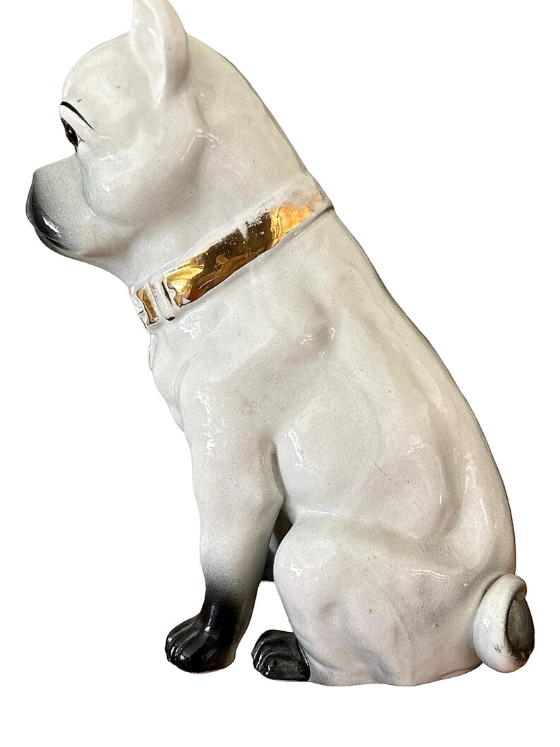 May include: A ceramic dog figurine in a sitting position. The dog is primarily white with black accents on the paws, nose, and ears. It has a gold-colored collar. The figurine has a glossy finish and is likely a decorative item.