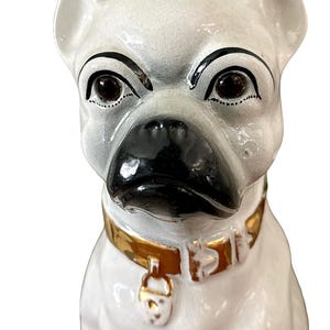 May include: A ceramic dog figurine with a white body, black nose, and dark eyes. The dog wears a gold-colored collar with a matching tag. The figurine has a slightly stern expression.