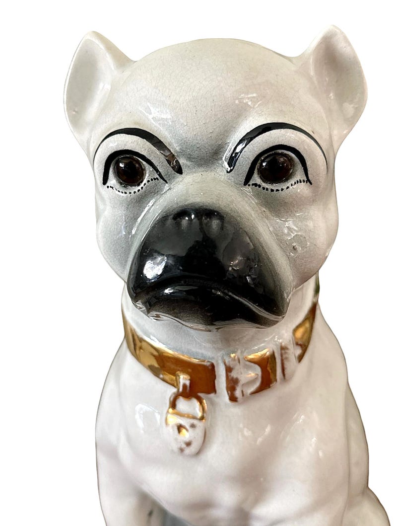 May include: A ceramic dog figurine with a white body, black nose, and dark eyes. The dog wears a gold-colored collar with a matching tag. The figurine has a slightly stern expression.