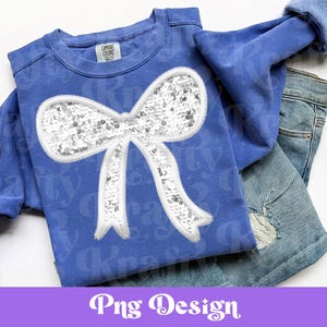 Puede incluir: Una sudadera azul con un gran diseño de lazo blanco con lentejuelas. La sudadera se combina con unos vaqueros. La imagen también incluye el texto "Png Design" en una pancarta morada en la parte inferior.