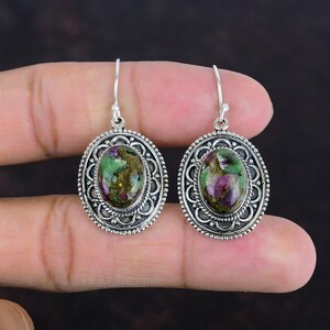 Könnte beinhalten: Ein Paar silberne Ohrringe mit ovalen, grün-rosa Edelsteineinsätzen. Die Ohrringe haben ein detailliertes, verziertes Design.