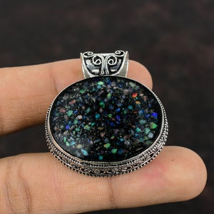 Pode incluir: Pingente oval com uma pedra escura e salpicada emoldurada em metal prateado. A pedra tem uma base preta com manchas azuis, verdes e vermelhas. O pingente tem uma borda ornamentada e uma argola decorativa.