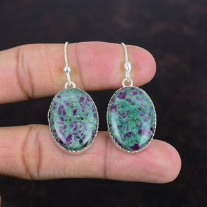 Könnte beinhalten: Ein Paar ovale Ohrringe mit grünen und violetten Edelsteinen, die in Silber gefasst sind. Die Edelsteine haben ein gesprenkeltes Aussehen, wobei Violett mit Grün kontrastiert. Die Ohrringe hängen an einfachen Silberhaken. Die Steine sind ca. 2 cm lang.