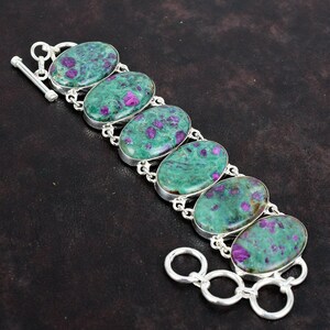 May include: A silver bracelet featuring six oval-shaped stones with a green base and purple flecks. The stones are set in silver and connected by silver links. The bracelet has a toggle clasp.