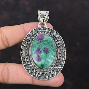 May include: Oval pendant with a green stone and pink inclusions, set in a silver frame. The pendant has a decorative border and a bail for a chain. The gemstone is the focal point of this jewelry piece, suitable for a necklace.