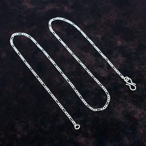 Puede incluir: Una cadena tipo fígaro de color plateado con un patrón repetitivo de tres eslabones circulares pequeños seguidos de un eslabón ovalado alargado. La cadena termina con un cierre de gancho simple en un extremo y una anilla circular en el otro.